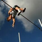 man in white tank top and black shorts hanging on black metal bar under blue sky